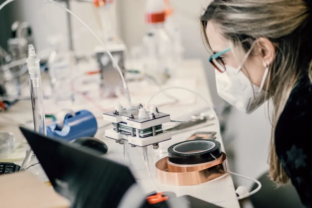 A student in a face mask examines a hardware prototype with tubes and copper tape on a lab bench