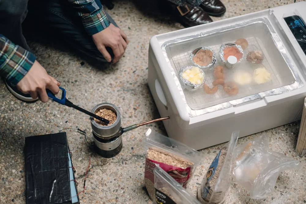 Hands working with tongs on a home-made smoker prototype next to a cooler box filled with food