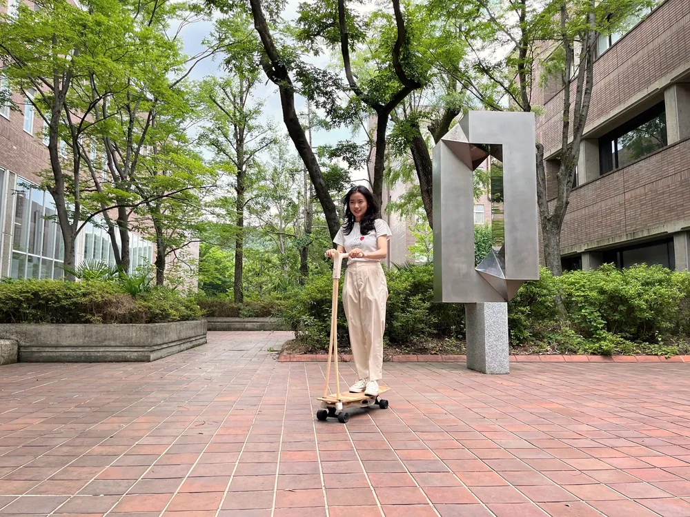 A student stands on a wooden board prototype with a tall handle in a tree-lined campus courtyard