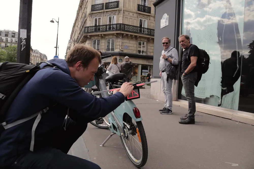 A student crouches beside a shared bike on a Paris street to test a prototype while two passers-by watch