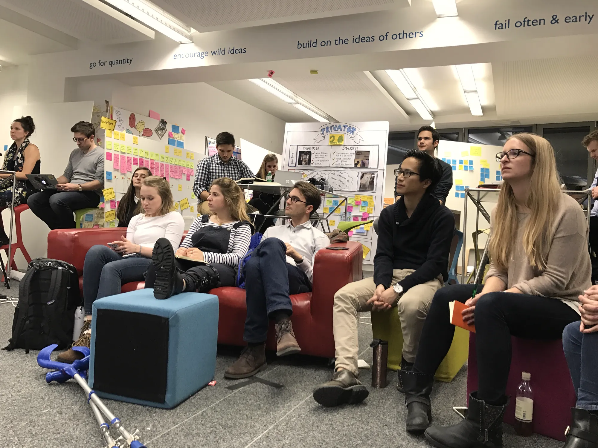 Students on sofas listen to a team pitch in front of walls covered with post-its