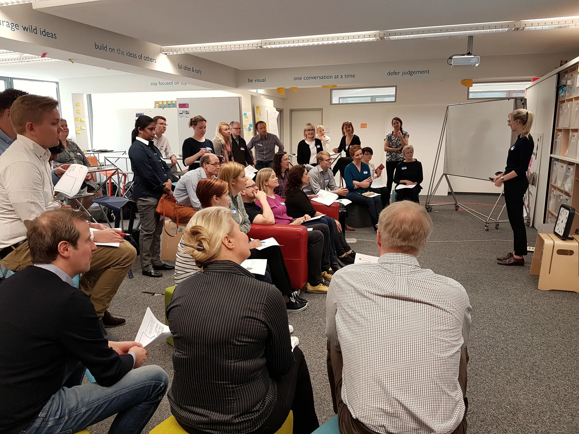 A facilitator briefs a seated group of participants in a workshop space with post-it walls