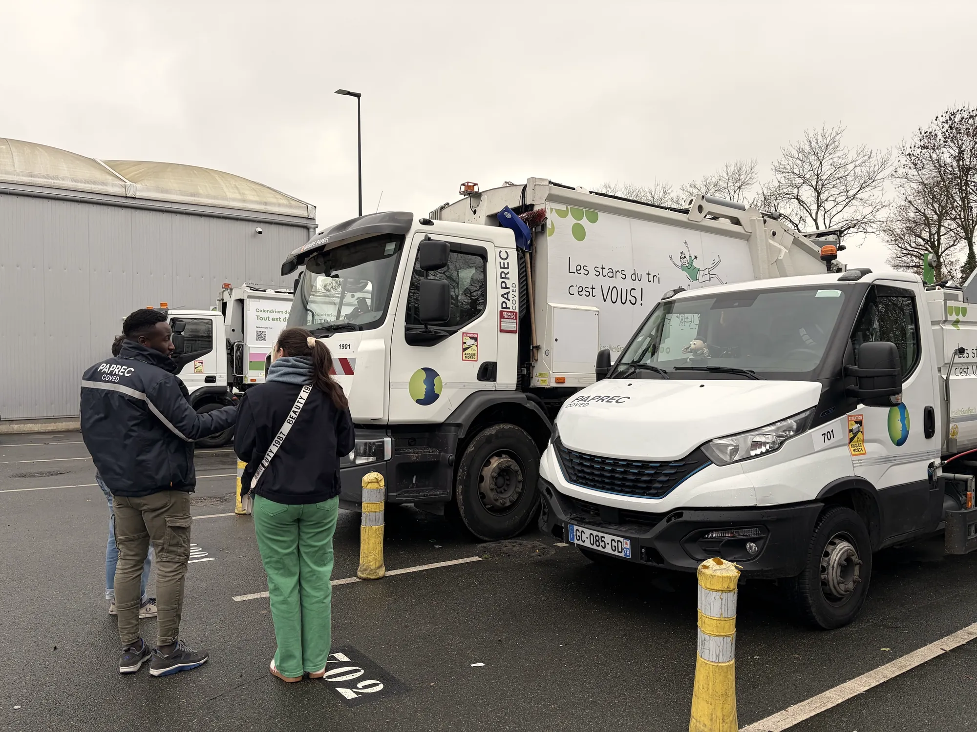 Students interview a waste-collection worker next to refuse trucks in a depot